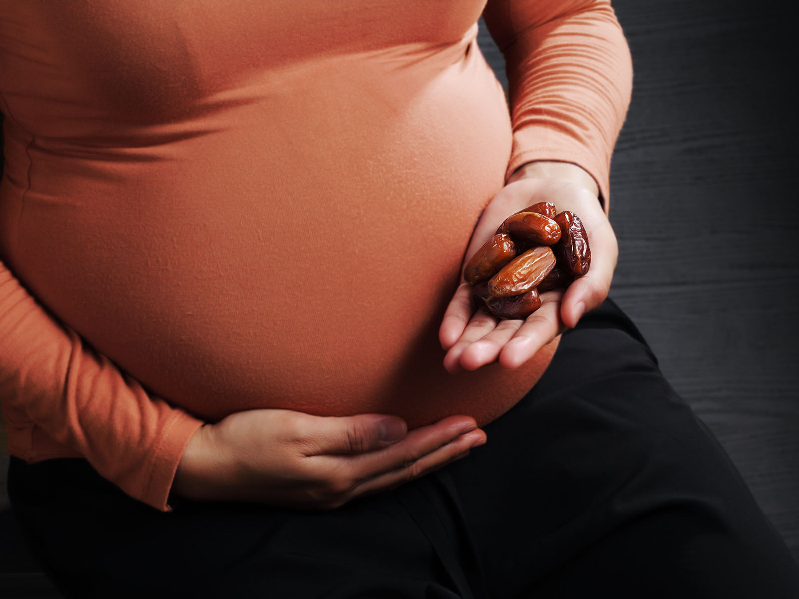 The image focuses on a pregnant woman, prominently displaying her rounded belly. She is wearing a long-sleeved, terracotta-colored shirt that closely conforms to the shape of her abdomen, emphasizing her pregnancy. Her hands are gently cradling her belly with one hand, while the other is outstretched, holding a small pile of dates. The dates are a rich, dark brown color and appear soft and slightly wrinkled, suggesting they are Medjool dates. The woman’s skin tone is warm and appears to be olive or tan. She is seated, wearing dark, possibly black, pants. The background is a dark, textured surface, resembling dark wood paneling, which provides a strong contrast to the warm tones of the woman’s clothing and skin. The lighting is soft and diffused, highlighting the curves of her body and the texture of the dates. The overall composition suggests a focus on health, nourishment, and the connection between pregnancy and natural foods. The image evokes a sense of calm and well-being, potentially promoting the benefits of dates during pregnancy, as they are known to be a source of energy and nutrients. The pose is intimate and protective, emphasizing the nurturing aspect of motherhood.